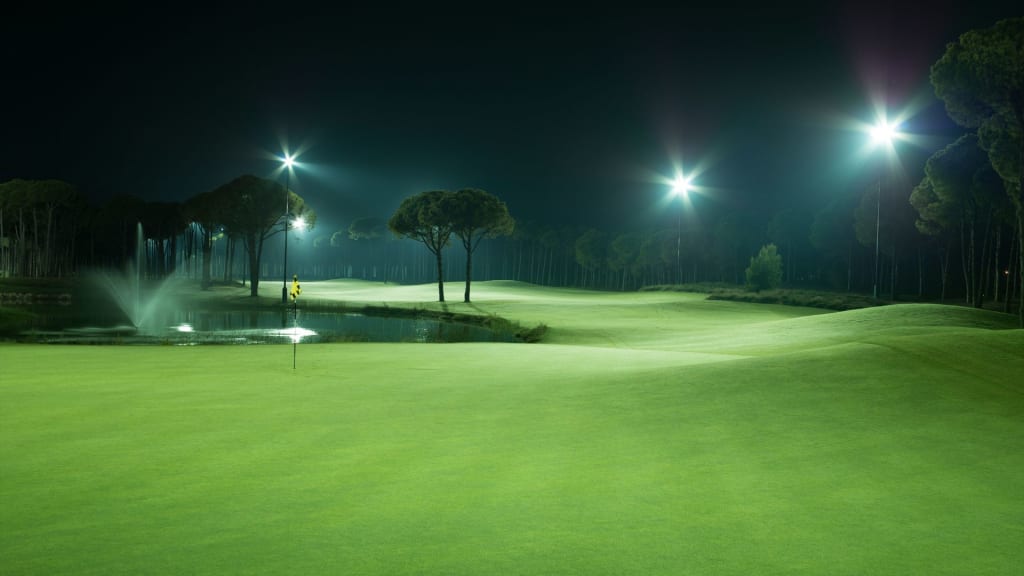 A golf course illuminated by bright floodlights at night, with a fountain in a pond near the green and trees lining the fairway.